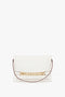 A white Nappa leather clutch handbag with a gold rectangular chain detail across the front and a thin brown strap on each side, embodying the elegance of a Chain Pouch Bag with Strap In White Leather by Victoria Beckham.