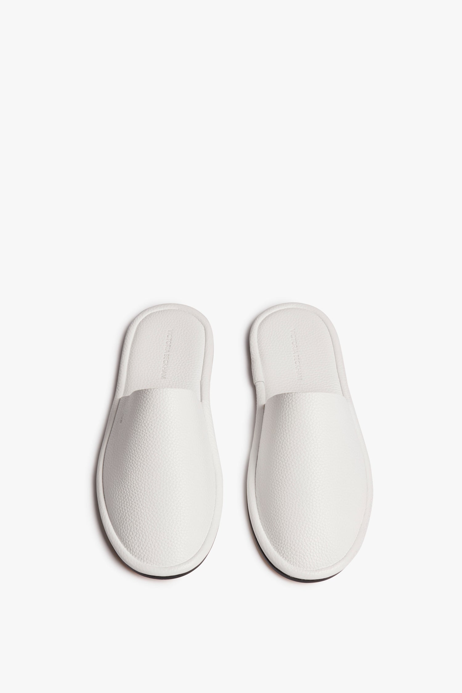 A pair of white, slip-on slippers with a textured surface are positioned side by side on a white background, embodying the chic simplicity of Victoria Beckham's Amelia Leather Mule in White design.