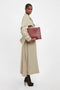 A person dressed in a beige trench coat and dark heels stands holding a Victoria Beckham Jumbo Chain Pouch Bag In Bordeaux with a detachable strap, crafted from grained leather, against a plain white background.