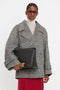 Person wearing a gray herringbone coat over a white turtleneck holds a Jumbo Chain Pouch Bag In Black Leather by Victoria Beckham featuring gold-tone chain detail and stands against a white background.