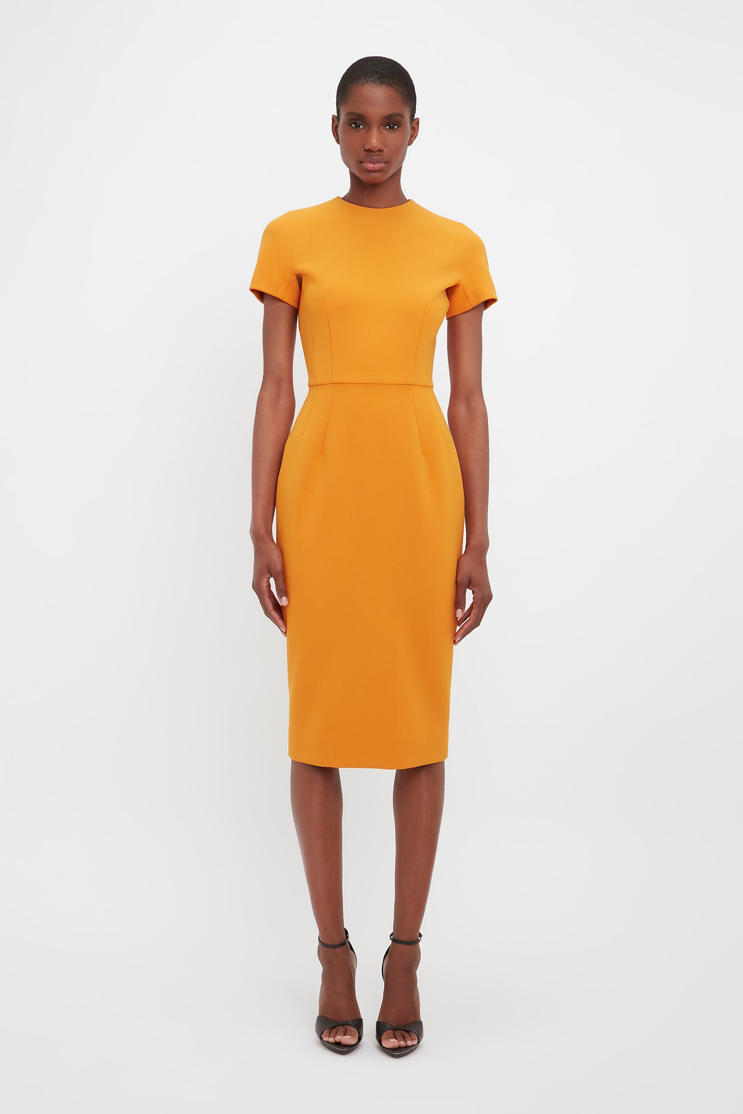 Model wearing the Dart Detailed Fitted T-shirt Dress in Mandarin for designer Victoria Beckham. This orange fitted pencil dress is perfect for workwear or wedding guest dressing. In a fitted style with short sleeves.