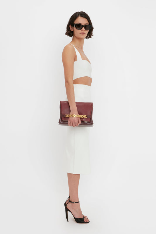 A woman in a Victoria Beckham Body Strap Bandeau Top In White and VB Body Fitted Midi Skirt stands sideways, holding a maroon handbag, wearing sunglasses, and high heels against a white background.