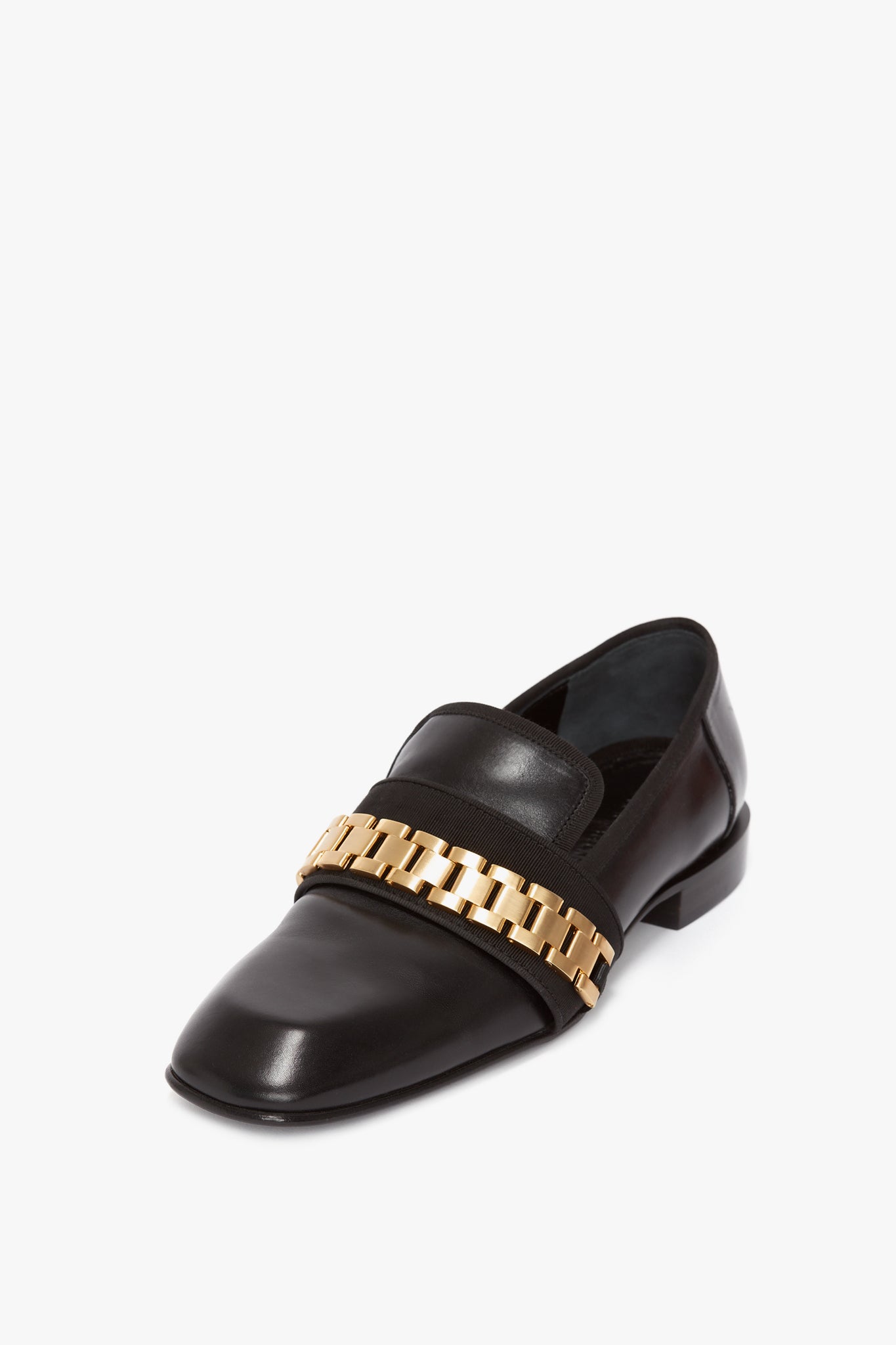The Mila Chain Loafer In Black by Victoria Beckham, a tuxedo shoe in shiny black patent leather, features a gold chain detail across the top and is elegantly displayed on a white background.