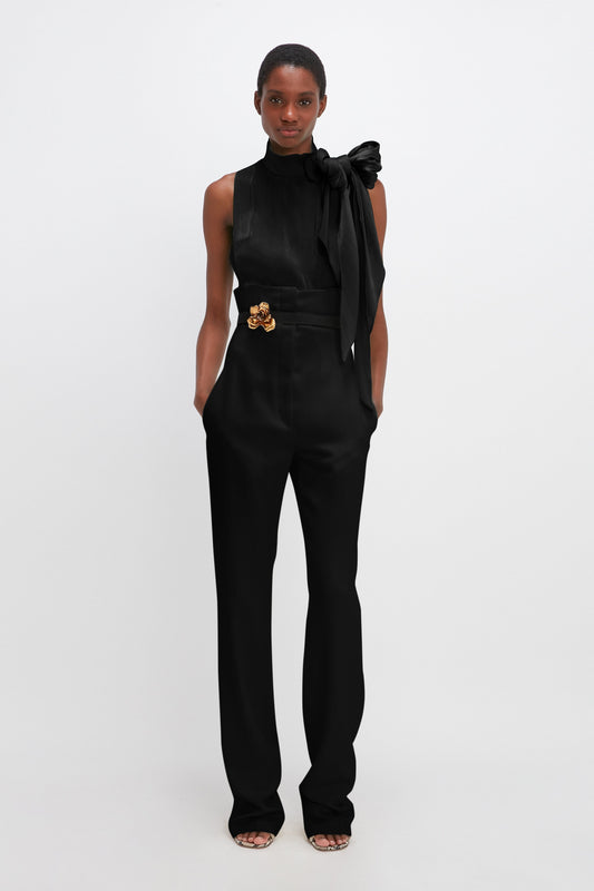 A woman stands against a grey background wearing a sleek black jumpsuit with a bow on the shoulder and a Victoria Beckham Exclusive Flower Belt in Black And Gold.